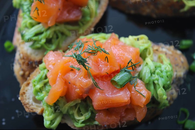 Gourmet avocado toast topped with smoked salmon, garnished with fresh dill and green onions on a black plate