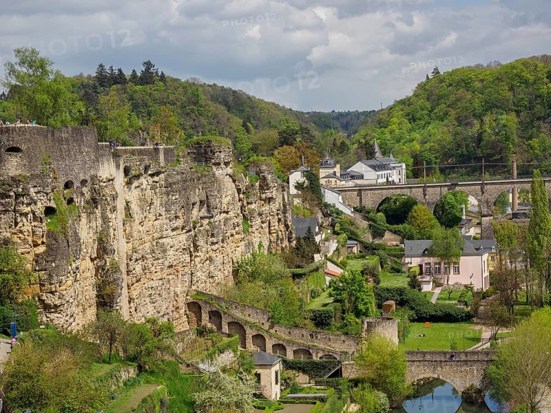 Rocky cliff landscape with a bridge, buildings and lush greenery around a river, overlooking a historic neighbourhood of a large seaside town with lots of greenery and old churches, Luxembourg, Europe