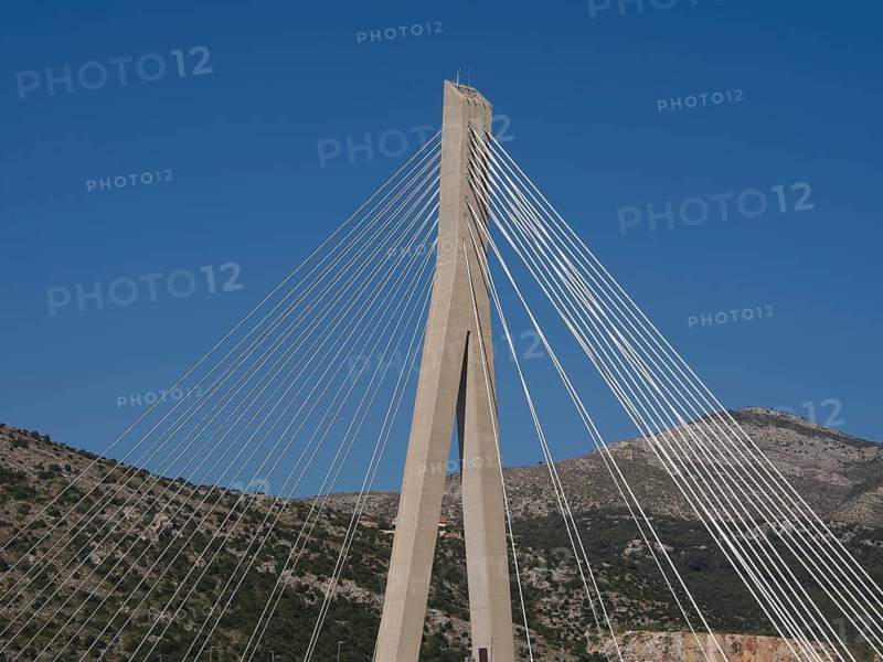 Modern suspension bridge with ropes in front of a clear blue sky and mountains, the old town of Dubrovnik with historic houses, churches, red roofs and fortress walls, dubrovnik, coratia