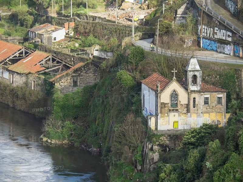 Small chapel and surrounding buildings on an overgrown, rocky cliff by a river with graffiti on walls, spring in the old town of Porto on the Douro with historic houses, Porto, portugal