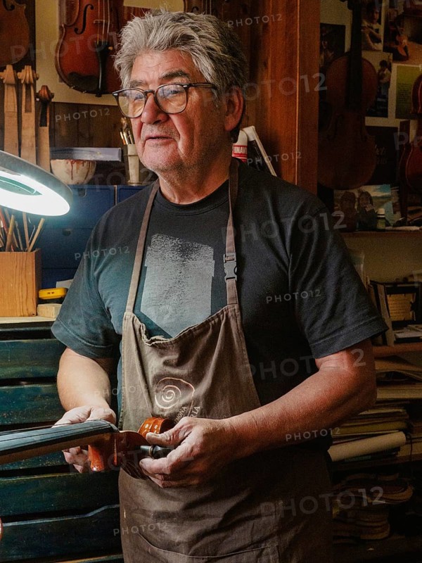 A craftsman wearing glasses and an apron works on a violin in a workshop, illuminated by a desk lamp