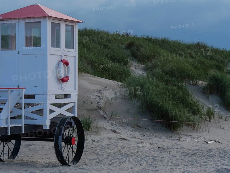 A white rescue tower with a red roof stands on a sandy beach in front of green dunes, Baltrum Germany