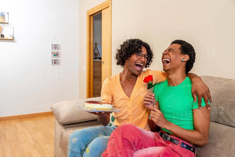Gay couple embracing celebrating love with a rose and cake sitting together in the sofa at home