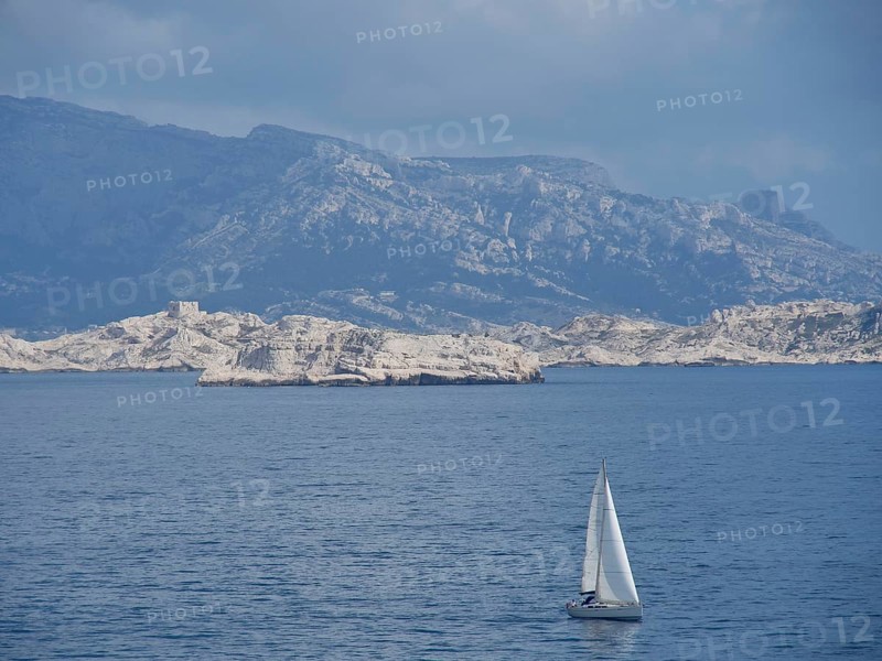 Sailboat sailing on a calm sea in front of a rocky mountain landscape and cloudy sky, Marseille on the Mediterranean Sea with the old cathedral and ships in the historic harbour, rocky coast on the Mediterranean Sea, marseille, france