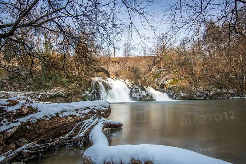 Beautiful landscape in winter with ice and snow. Waterfall of the river Elz near the castle Pyrmont, the country inn Pyrmonter Muehle directly at a lake. A historic bridge in Rhineland-Palatinate, Germany, Europe