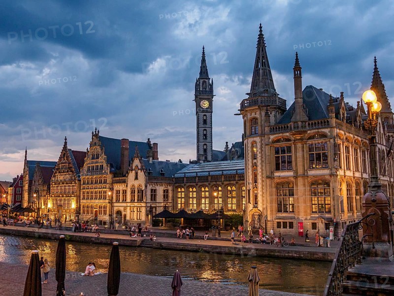 Gothic buildings with illuminated windows and people walking along a canal under a cloudy sky, historic buildings with towers and bridges by a river at blue hour, gent, belgium