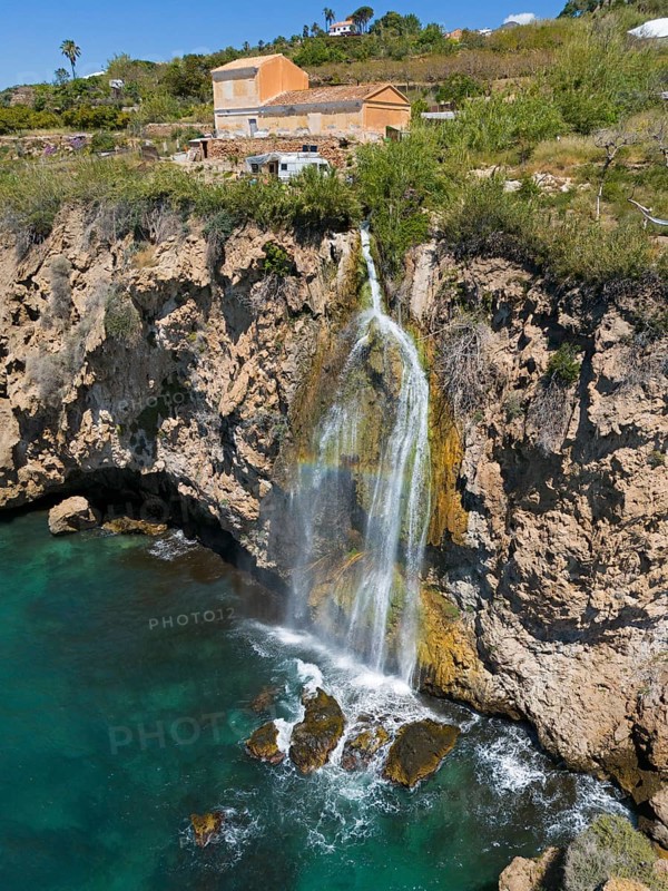 A picturesque waterfall falls from a cliff into turquoise water with houses in the background, a rocky, natural environment, aerial view, Cascada Grande de Maro, Nerja, Malaga, Andalucia, Spain, Europe