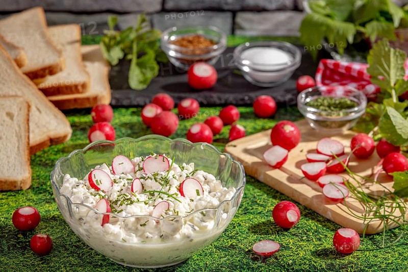 Healthy spread of radishes, cottage cheese and herbs surrounded by ingredients and slices of bread on a grassy area