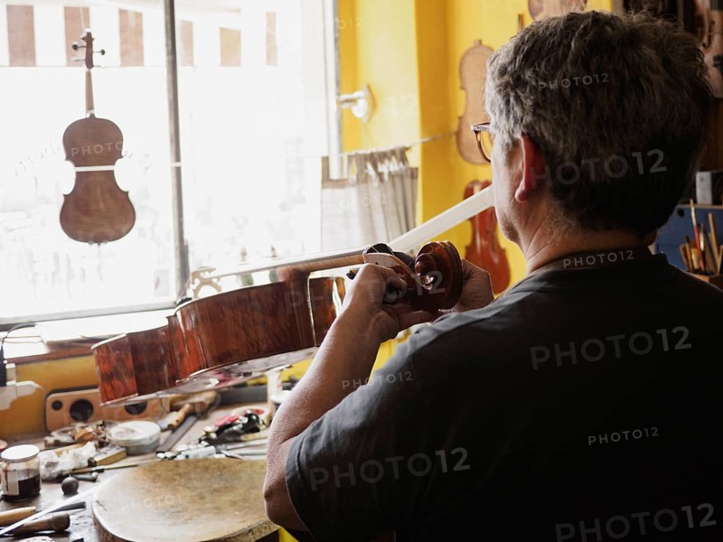 From behind, a luthier works intently on a cello in a well-organized workshop with natural light