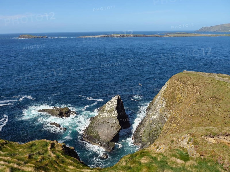 Impressive cliff landscape with rough waves crashing against rocks, under a blue sky, green meadows by the sea with stony shores and ruins, Lerwick, shetland