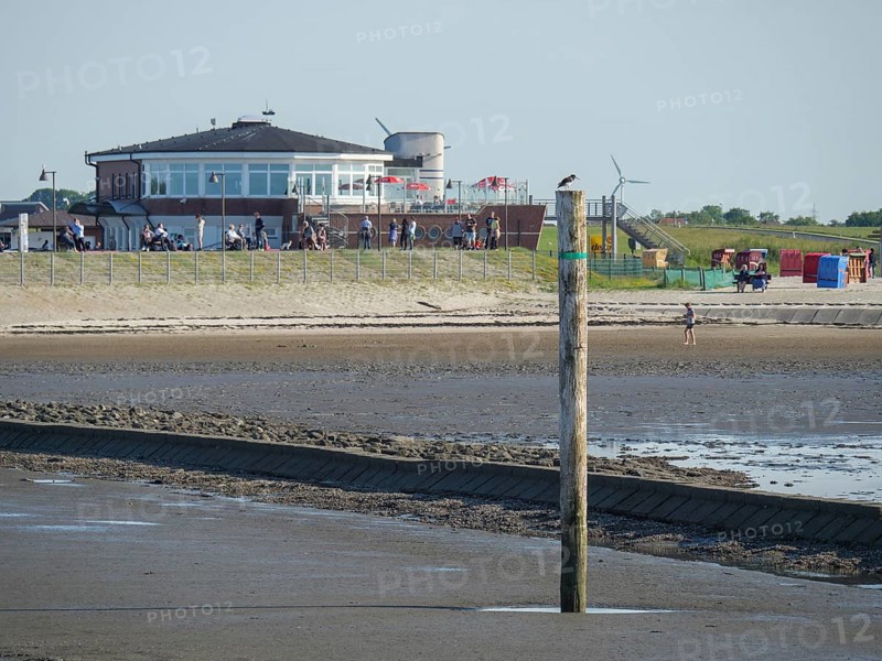 A pole on the beach with water in front of it, in the background a modern building and beach chairs, people on the beach, Baltrum Germany