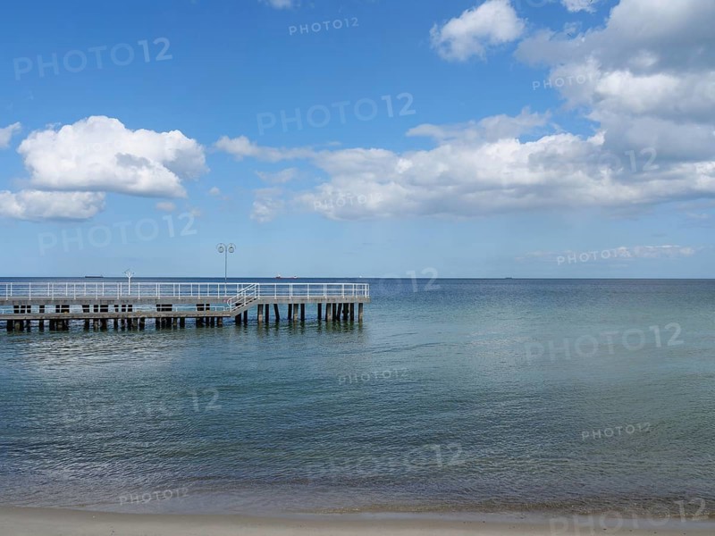 A jetty juts out over the calm waters of the sea, the sandy beach leads into the distance under a blue sky with clouds, Green trees on a Baltic beach with clear water, Sopot, poland