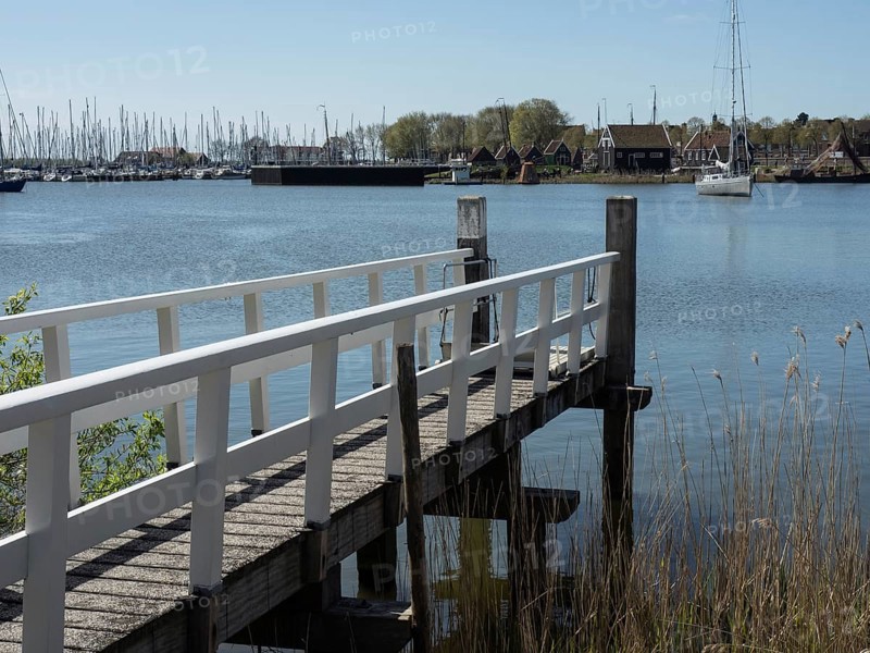 A wooden jetty leads into a lake, boats are moored on the shore, the surroundings appear calm and peaceful, Enkhuizen, Nirderlande
