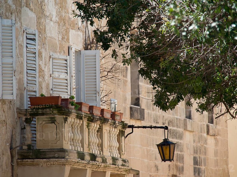 Mediterranean balcony with plants and open shutters, next to it a lantern on an old stone wall, Historic buildings with beautiful windows, balconies and small towers, Mdina, Malta, Europe