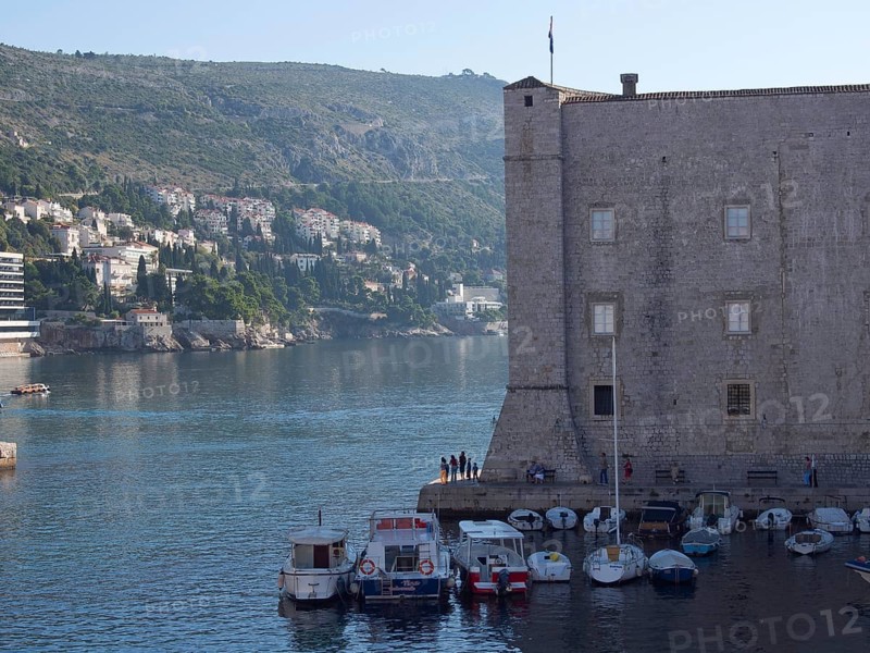 Stone building at the edge of a harbour with boats in the water, city and mountains in the background, the old town of Dubrovnik with historic houses, churches, red roofs and fortress walls, dubrovnik, coratia