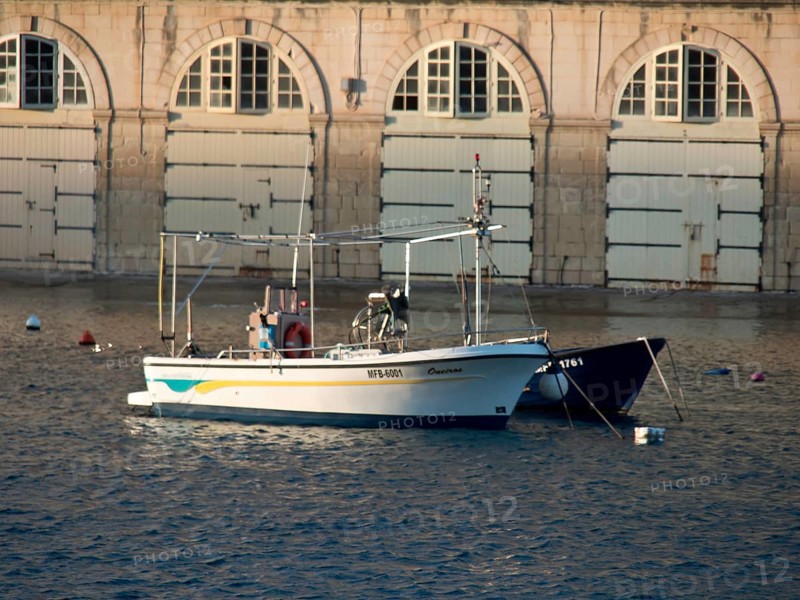 A boat in the harbour in front of a building with stone walls and many windows, small boats in the harbour in front of historic buildings in the evening light, Valetta, Malta, Europe