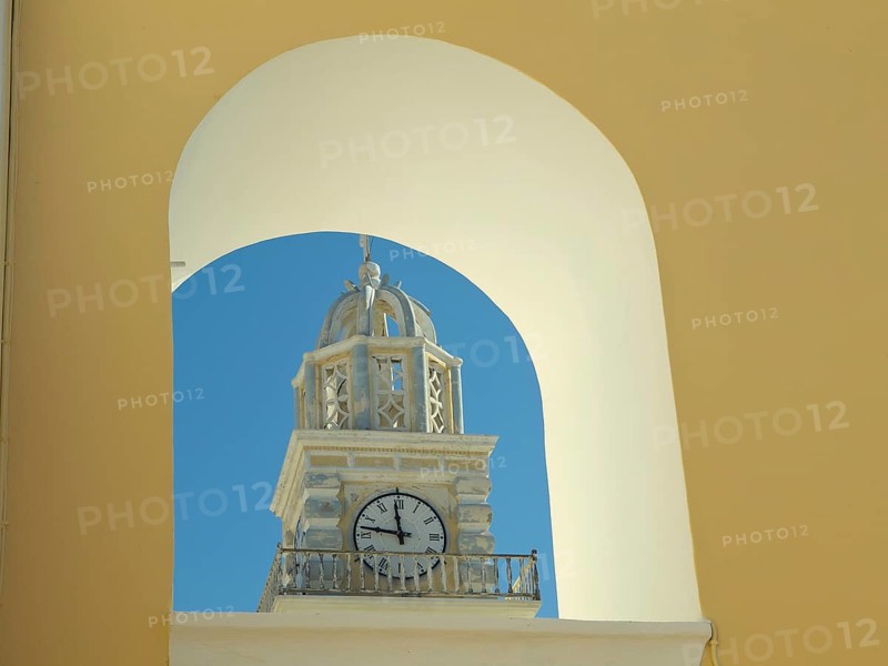 The clock of a church tower seen through a yellow arch, contrasted against the blue sky in Santorini, The volcanic island of Santorini with blue and white houses and churches, flowers and the deep blue sea, Santorini, Greece, Europe