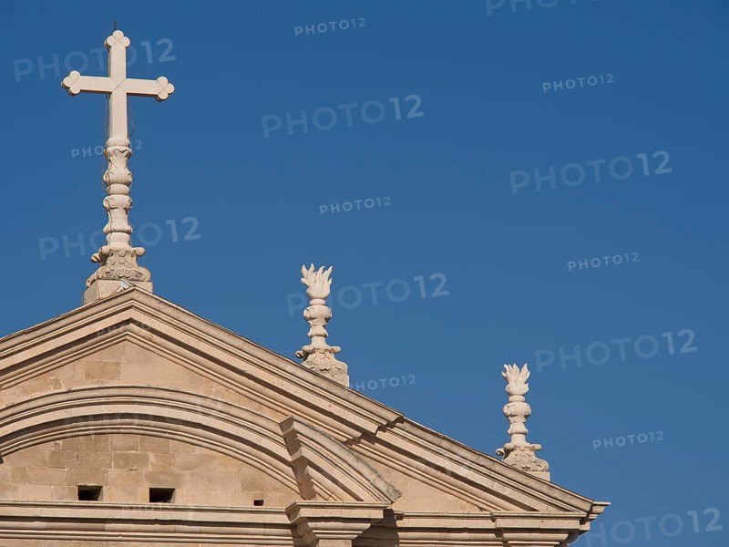 Top of a church with a cross and classical style elements in front of a cloudless blue sky, the old town of Dubrovnik with historic houses, churches, red roofs and fortress walls, dubrovnik, coratia