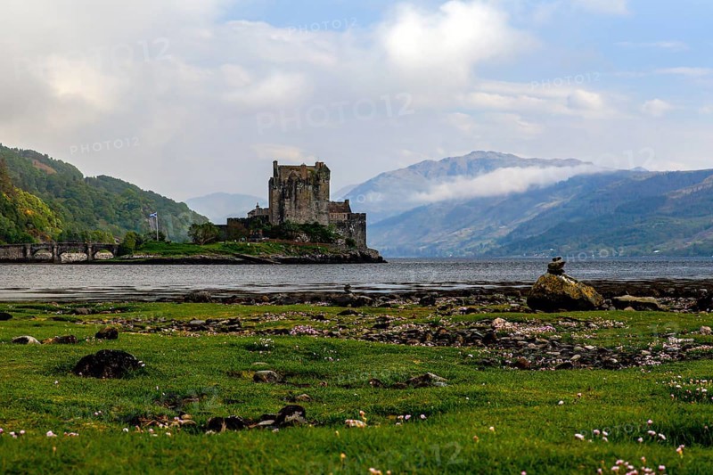 A castle on the lakeshore with mountains and clouds in the background, surrounded by green meadows and stones