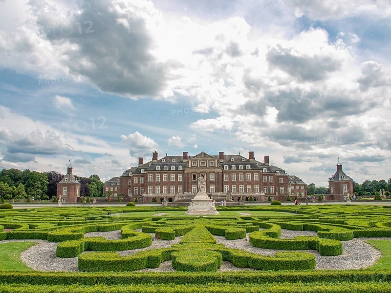 A large historic castle surrounded by a symmetrical garden with manicured hedges under a cloudy sky, historic castle with a green park with sculptures and paths, nordkirchen, germany