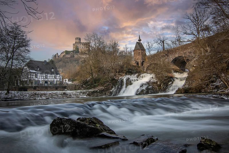 Beautiful landscape in winter with ice and snow. Waterfall of the river Elz near the castle Pyrmont, the country inn Pyrmonter Muehle directly at a lake. A historic bridge in Rhineland-Palatinate, Germany, Europe