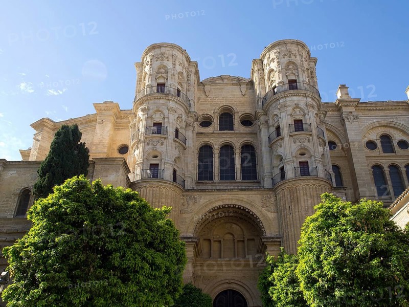 A historic cathedral with two towers and stone facade surrounded by trees on a sunny day, The city of Malaga on the Mediterranean Sea with the historic fortress and palm trees, Malaga, spain