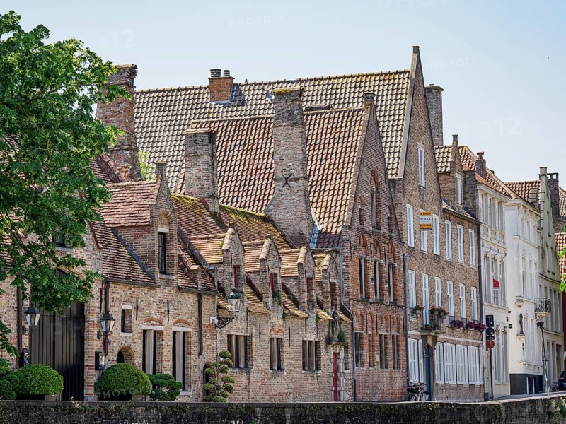 Medieval brick buildings with several chimneys along a canal, surrounded by trees, historic houses and churches with towers on a river in Belgium, Bruges, Belgium, Europe