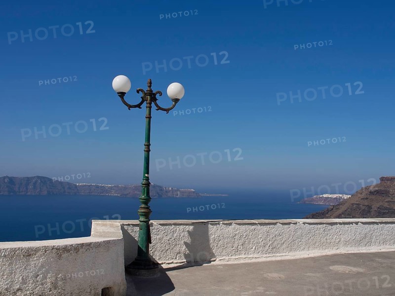Street lamp on a white terrace with a view of the sea and mountains in the background, The volcanic island of Santorini with blue and white houses and churches, flowers and palm trees on a volcanic island, santorini, greece
