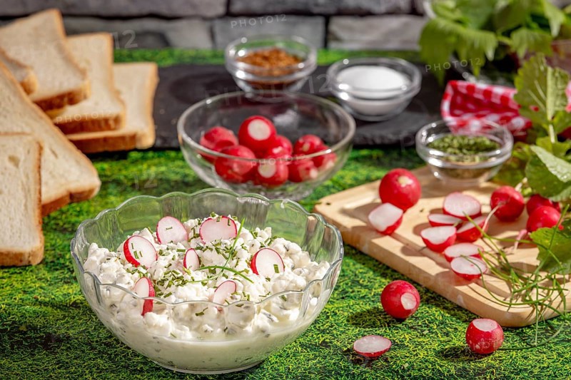 Fresh spread of radishes, cottage cheese and spices surrounded by ingredients and slices of bread on a grassy area
