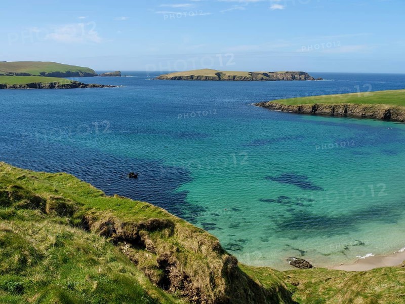 Turquoise waters meet rocky cliffs and green grasslands under a blue sky, Green meadows by the sea with rocky shores and ruins, Lerwick, shetland