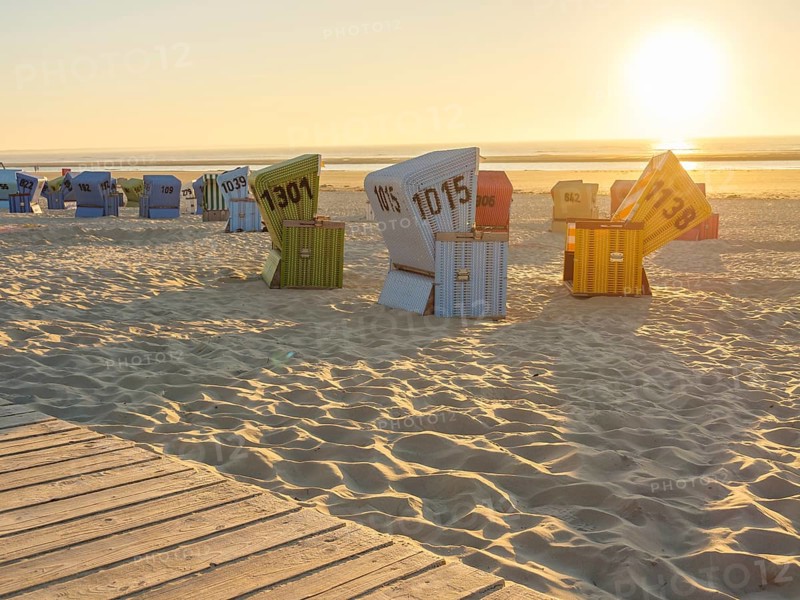 Several colourful beach chairs standing in order in the sand while the sun illuminates the horizon, a wooden path leads to the beach, setting sun on the North Sea beach with warm light over colourful beach chairs, langeoog, germany