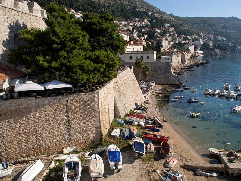 Boats moored on the shore next to a city wall, the sun shining brightly and the clear sky adding to the summer atmosphere, the old town of Dubrovnik with its historic houses, churches, red roofs and fortress walls, dubrovnik, coratia