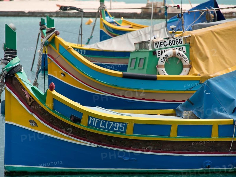 Close up of colourful fishing boats in the water, with bright colours and ropes, many colourful fishing boats in a small harbour against a historical backdrop, Marsaxlokk, malta