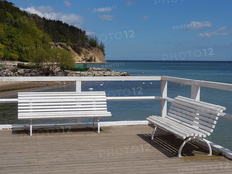White benches on a jetty, behind them the sea and a wooded cliff, spring on the Baltic coast with green fishing boats on the beach and green trees on the shore, Sopot, Poland, Europe