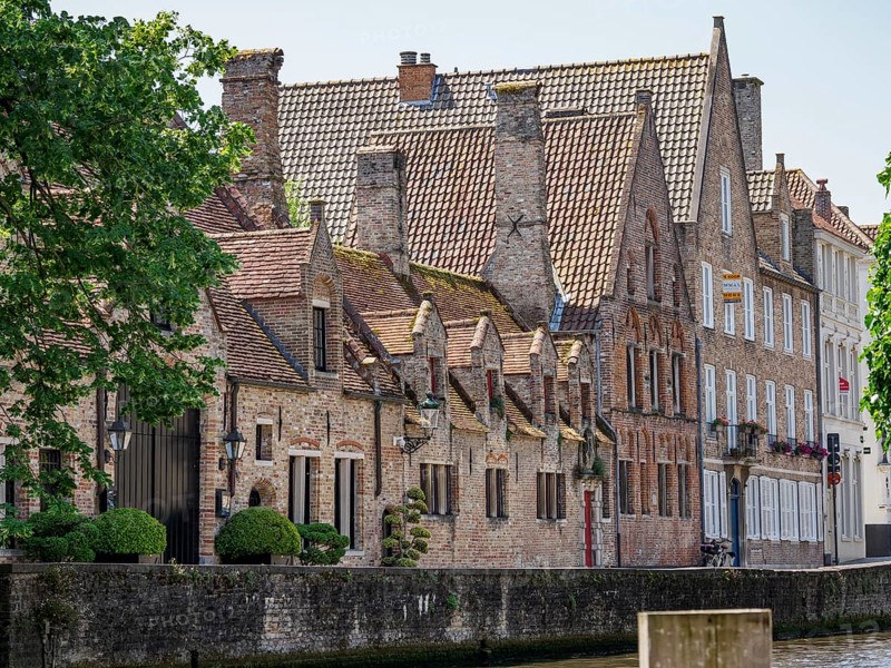Canal with neighbouring old buildings and numerous chimneys, flanked by green trees, historic houses and churches with towers on a river in Belgium, Bruges, Belgium, Europe