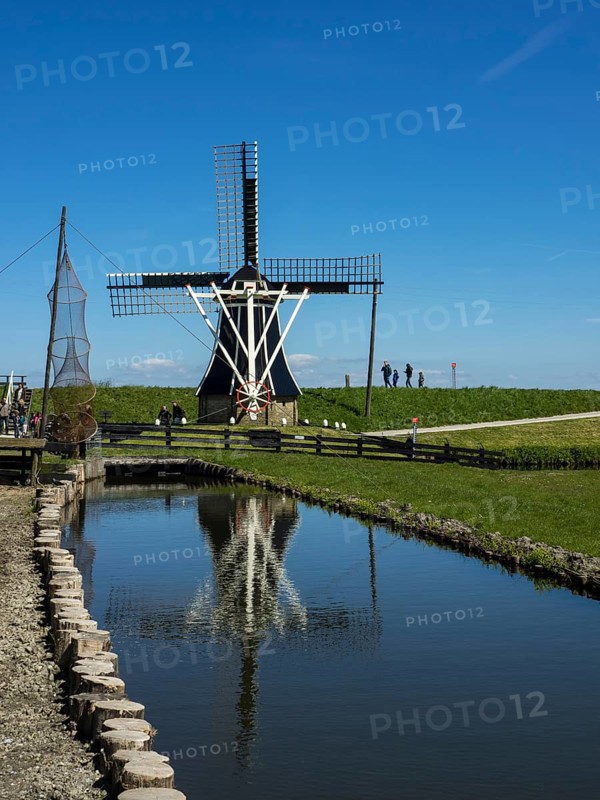 Large windmill with its reflection in a pond, surrounded by people, under a clear blue sky, Enkhuizen, Nirderlande