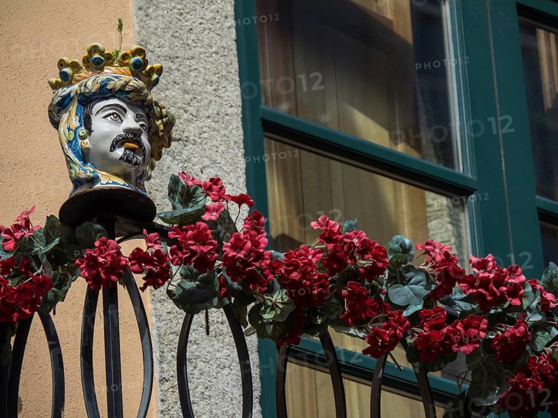 Ceramic bust on a balcony with red flowers in front of a window on a rustic facade, The old town of Porto on the Douro River with colourful houses and a large bridge, Porto, Portugal, Europe