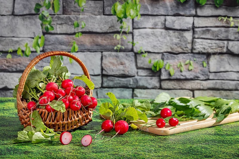A basket of fresh radishes and a chopping board with herbs on a grassy area in front of a stone wall