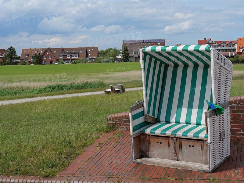 A green and white beach chair stands on a paved square with houses in the background, Baltrum Germany