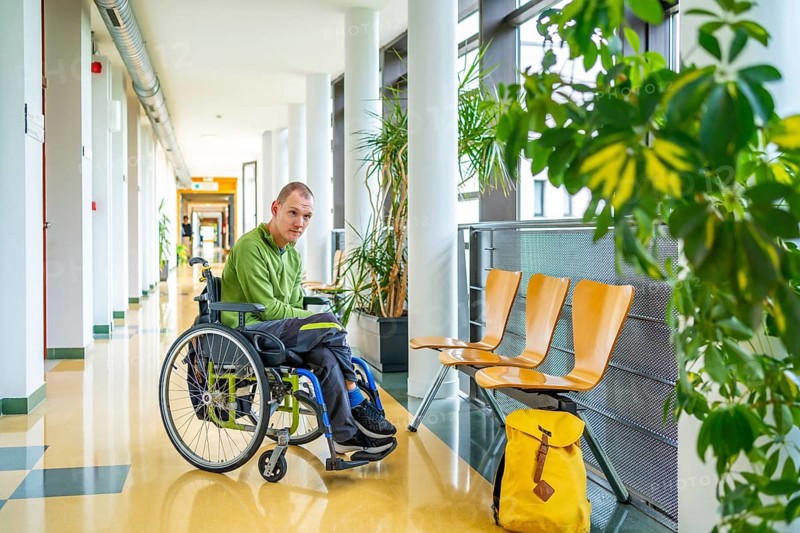 Man with cerebral palsy in wheelchair in the university waiting in the corridor next to school bag