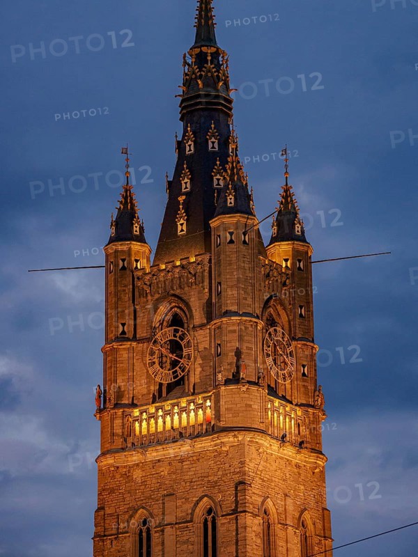 Illuminated clock tower with gothic elements in the evening, blue hour in a medieval town with historic buildings with towers and bridges on a river, gent, belgium