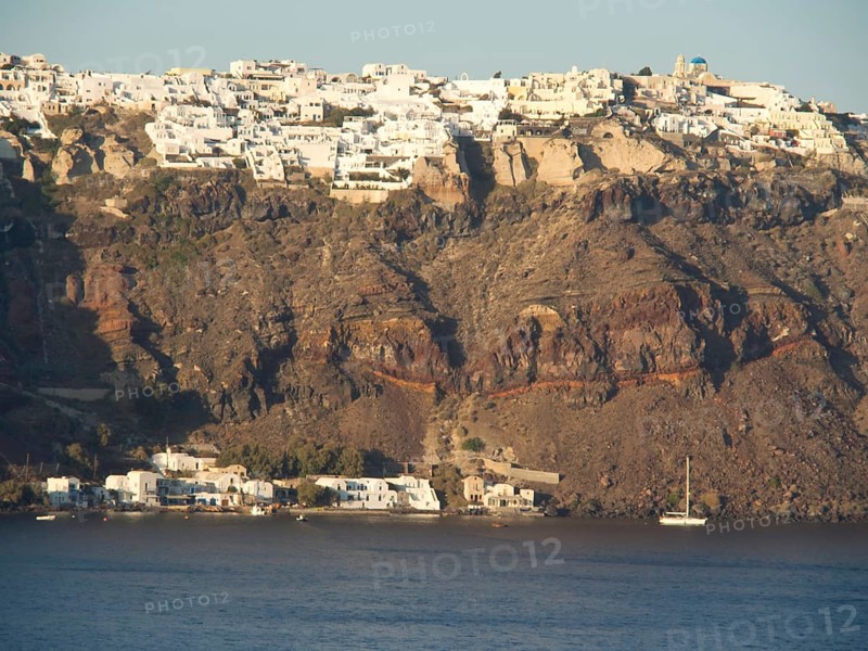 A cluster of white buildings on a cliff dramatically jutting out over the calm sea, brown volcanic island in the blue sea with white houses, santorini, greece