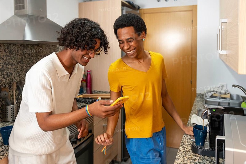 Gay couple smiling while using phone preparing coffee at home in the morning