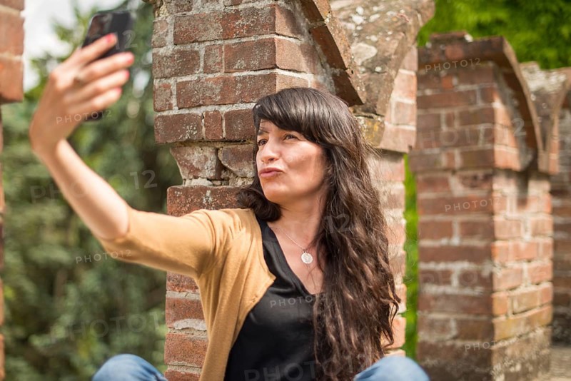 Young hispanic woman in casual clothing focused on conversation with smartphone in a peaceful garden with blurred brickwork in the foreground