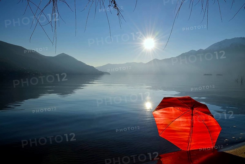 Red Umbrella Leaning on the Sand Beach on Alpine Lake Maggiore with Mountain and Sunbeam and Brissago Islands in a Sunny Day in Ascona, Ticino, Switzerland, Europe