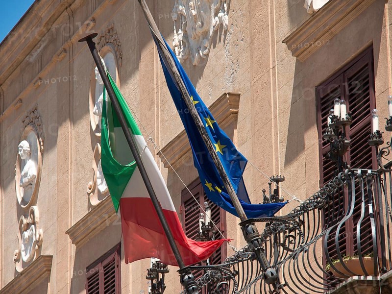 A building with an Italian and European flag, restrained historical architecture, palermo in sicily with an impressive cathedral, monuments and old houses with balconies, palm trees and flowers in italy, palermo, sicily