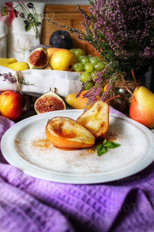 A rustic still life of fruits including pears, figs, and grapes, accented with spices and a bouquet of purple flowers on a purple cloth