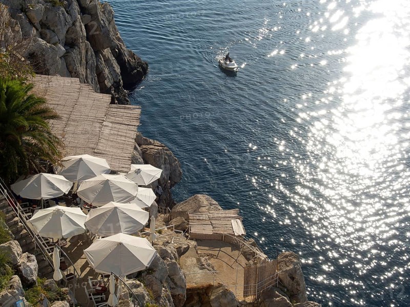 View of the sea from the rock, with white parasols and a boat on the water, reflecting the sun's rays, the old town of Dubrovnik with historic houses, churches, red roofs and fortress walls, dubrovnik, coratia
