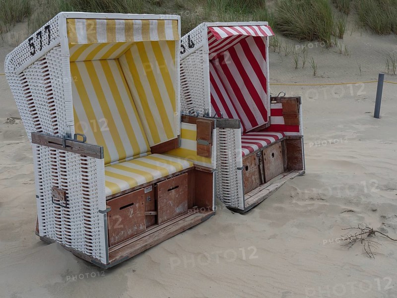 Two beach chairs, one yellow and one red, on the sandy beach in front of the dunes, the north sea island baltrum with grassy dune, colourful beach chairs on the beach and clouds over the sea, Baltrum Germany