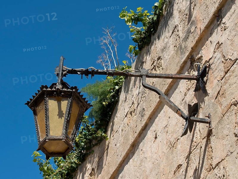 An old metal lantern on a stone wall, overgrown with plants under a blue sky, the town of mdina on the island of malta with historic houses, colourful balconies, magnificent churches, mdina, Malta, Europe
